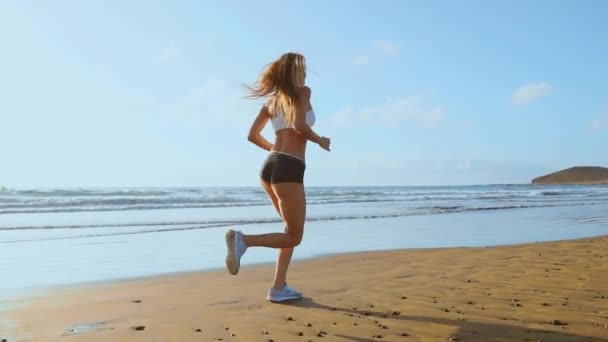 穿着运动短裤和 t恤衫的美丽女子, 在沙滩上奔跑, 白色的沙子和蓝色的海水在岛上缓慢运动。波浪和沙丘陵的背面赢得了 — 图库视频影像