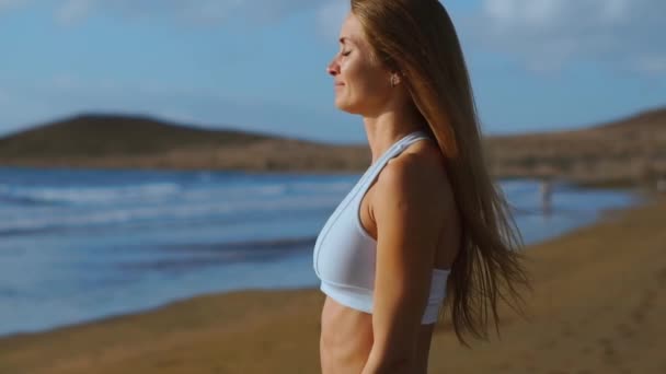 Uma mulher de roupas esportivas chega ao oceano e olha para a distância tocando seu cabelo. SLOW MOTION STEADICAM — Vídeo de Stock