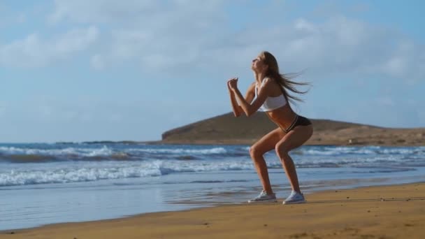 Fitness mujer joven ejercitando el núcleo y los glúteos con entrenamiento de peso corporal haciendo ejercicios en cuclillas en la playa. chica deportiva en cuclillas piernas como parte de una vida activa y en forma. sesión de stedicam — Vídeos de Stock