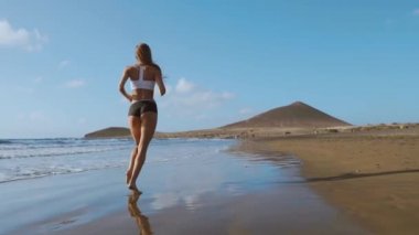 Plajda koşan fitness koşucusu kadın telefon kılıfı kolluk bandıyla müzik dinliyor. Spor atleti yaz güneşi altında çıplak ayakla kardiyo antrenmanı yapıyor..