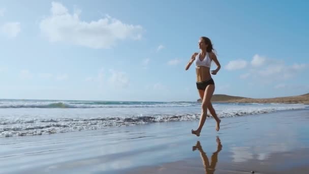 Бегущая по пляжу женщина слушает музыку с телефоном, спортивными арматурой и лямкой. Спортивный спортсмен тренирует кардио босиком с решимостью под летним солнцем. — стоковое видео