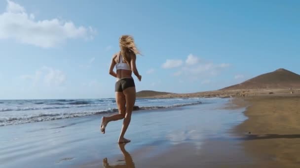 Bella donna sportiva che corre lungo la bellissima spiaggia di sabbia, stile di vita sano, godendo di una vacanza estiva attiva vicino al mare. SLOW MOTION STEADICAM . — Video Stock