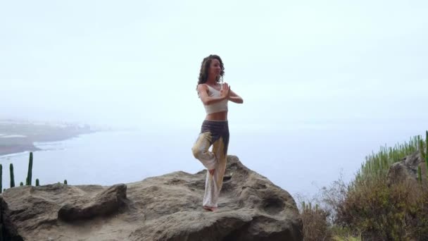 Mujer joven haciendo yoga en las montañas de pie en una pierna con vistas al océano — Vídeos de Stock