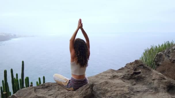 Jonge vrouw doen yoga in de bergen op een eiland met uitzicht op de oceaan zittend op een rots op de top van een berg mediteren in de lotuspositie — Stockvideo