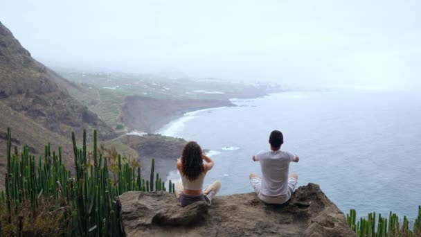 Un hombre y una mujer sentados en la cima de una montaña mirando al océano sentados en una piedra meditando levantando las manos y realizando un respiro relajante. Islas Canarias — Vídeo de stock