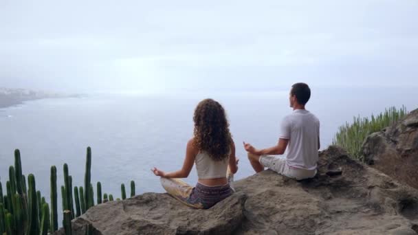 Un hombre y una mujer sentados en la cima de una montaña mirando al océano sentados en una piedra meditando en una posición de Loto. La vista desde atrás. Islas Canarias — Vídeo de stock