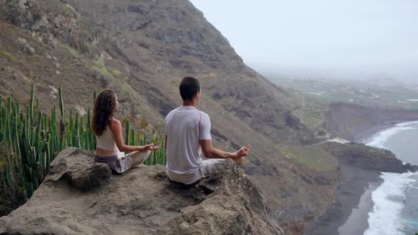 Un hombre y una mujer sentados en la cima de una montaña mirando al océano sentados en una piedra meditando en una posición de Loto. La vista desde atrás. Islas Canarias — Vídeo de stock