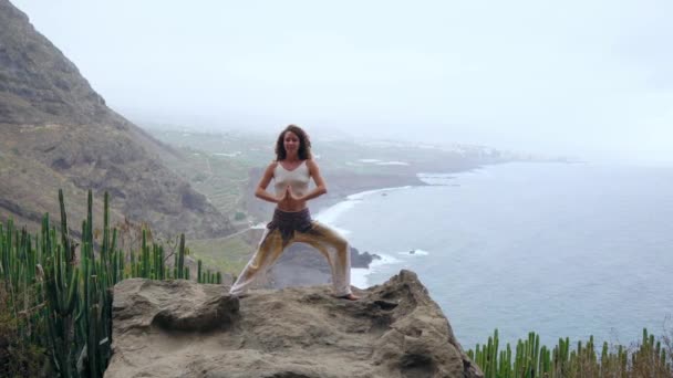 Vrouw in yoga warrior pose op de Oceaan, strand en rock bergen mediteren. Motivatie en inspirerende pasvorm en oefenen. Gezonde levensstijl buiten in de natuur, fitness concept. — Stockvideo
