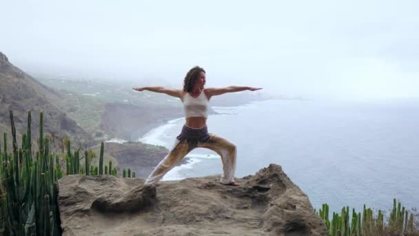 Woman meditating in yoga warrior pose at the ocean, beach and rock mountains. Motivation and inspirational fit and exercising. Healthy lifestyle outdoors in nature, fitness concept. — Stock Video