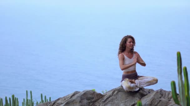 Jeune femme faisant du yoga dans les montagnes sur une île surplombant l'océan assis sur un rocher au sommet d'une montagne méditant en position Lotus — Video