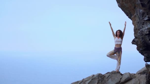 Jonge vrouw doet yoga op een rotsachtige kust bij zonsondergang. Het concept van een gezonde levensstijl. Harmonie. Mens en natuur. De achtergrond van de blauwe oceaan. — Stockvideo