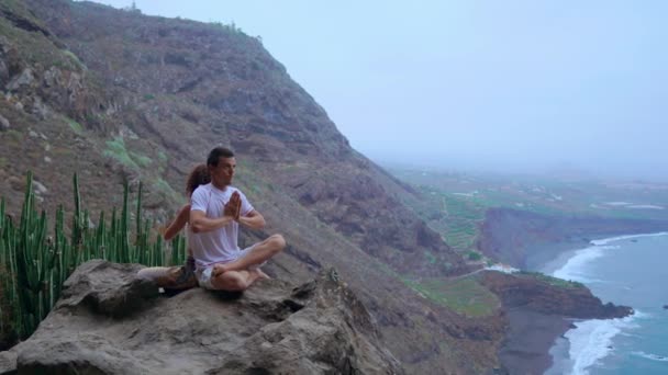 Man en vrouw zit op de top van een berg op een rots rug aan rug mediteren en het doen van yoga op de achtergrond van de Oceaan. — Stockvideo