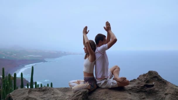 Man and woman sitting on top of a mountain on a rock back to back meditate and do yoga on the background of the ocean. — Stock Video