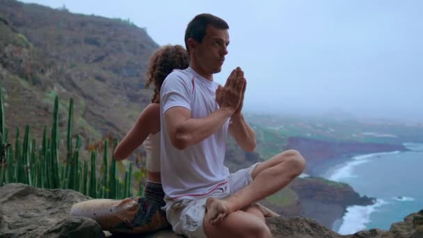 Mannen och kvinnan sitter ovanpå ett berg på en sten rygg mot rygg meditera och göra yoga på bakgrunden av havet. — Stockvideo