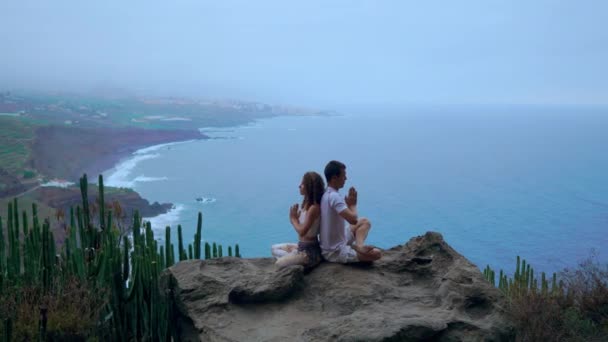 Mannen och kvinnan sitter ovanpå ett berg på en sten rygg mot rygg meditera och göra yoga på bakgrunden av havet. — Stockvideo