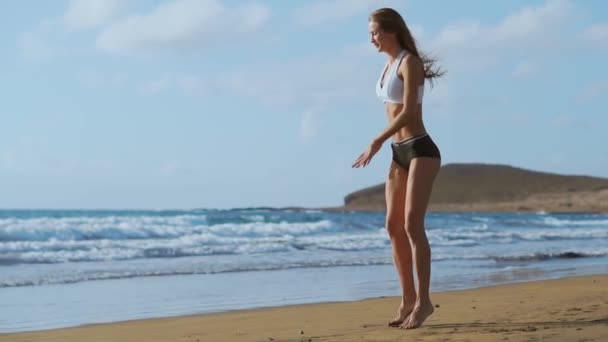 Menina em shorts sportswear e t-shirt executa saltos com agachamentos na praia perto do oceano nas Ilhas Canárias. Estilo de vida saudável e fitness nas férias. Corpo bonito e saudável — Vídeo de Stock