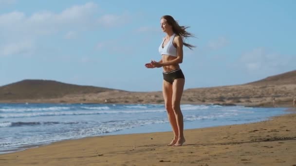 Chica en pantalones cortos de ropa deportiva y camiseta realiza saltos con sentadillas y palmadas en la playa cerca del océano en las Islas Canarias. Vida sana y forma física en vacaciones. Hermoso y saludable cuerpo — Vídeos de Stock