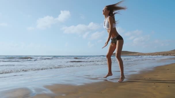 Menina em shorts sportswear e t-shirt executa saltos com agachamentos e palmas na praia perto do oceano nas Ilhas Canárias. Estilo de vida saudável e fitness nas férias. Corpo bonito e saudável — Vídeo de Stock