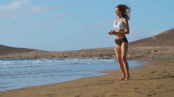 Menina em shorts sportswear e t-shirt executa saltos com agachamentos na praia perto do oceano nas Ilhas Canárias. Estilo de vida saudável e fitness nas férias. Corpo bonito e saudável — Vídeo de Stock