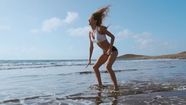 Girl in sportswear shorts and t-shirt performs jumps with squats on the beach near the ocean in the Canary Islands. Healthy lifestyle and fitness on vacation. Beautiful and healthy body — Stock Video