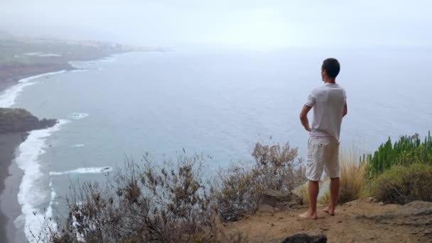 Чоловік, що стоїть на краю скелі з видом на океан, піднімає руки і вдихає морське повітря під час йоги — стокове відео