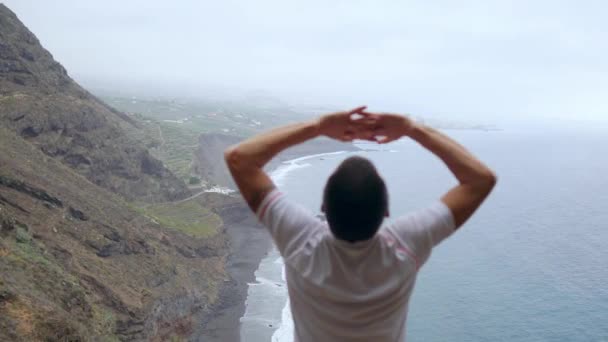 En man som står på kanten av en klippa med utsikt över havet höjer händerna upp och andas in havsluften under yoga — Stockvideo