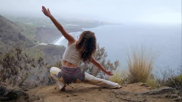 Una mujer sentada en el borde de un acantilado en una pose de guerra con vistas al océano levanta las manos e inhala el aire del mar mientras hace yoga — Vídeos de Stock