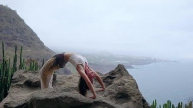 Kızı pratik yoga mavi gökyüzü ve masmavi denizin karşı buzlu. Kadın bir köprü duruş bir taş üzerinde duruyor.