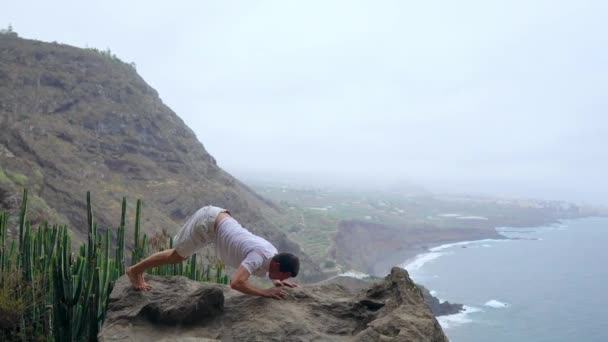 L'uomo in piedi sul bordo di una scogliera in posa del cane con vista sull'oceano, respirare l'aria di mare durante un viaggio yoga attraverso le isole — Video Stock