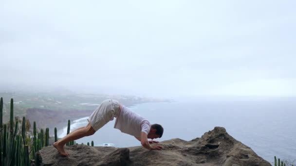 L'homme debout sur le bord d'une falaise dans la pose du chien avec vue sur l'océan, respirer l'air de la mer pendant un voyage de yoga à travers les îles — Video