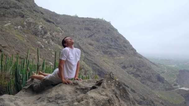 Чоловік, що стоїть на краю скелі в позу з собаки з видом на океан, вдихаючи морського повітря під час йога подорож по островах — стокове відео