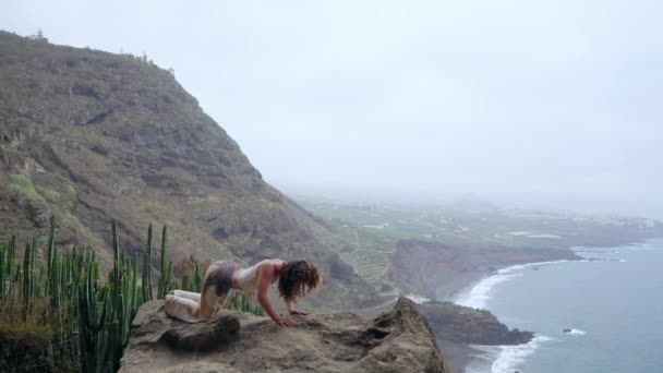 Jovem mulher caucasiana realizando postura de cão virada para cima ao ar livreA mulher sentada na borda de um penhasco na pose do cão com vista para o oceano, respirar o ar do mar durante uma viagem de ioga — Vídeo de Stock