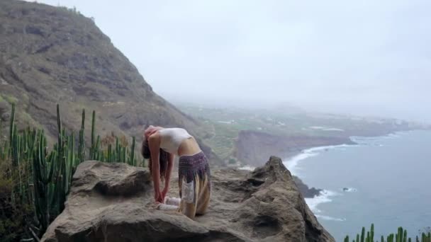 Mädchen praktiziert Yoga auf den Felsen vor dem blauen Himmel und dem azurblauen Meer. Frau steht auf einem Stein in Brückenhaltung. — Stockvideo