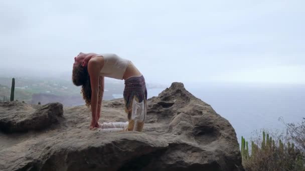 Mädchen praktiziert Yoga auf den Felsen vor dem blauen Himmel und dem azurblauen Meer. Frau steht auf einem Stein in Brückenhaltung. — Stockvideo