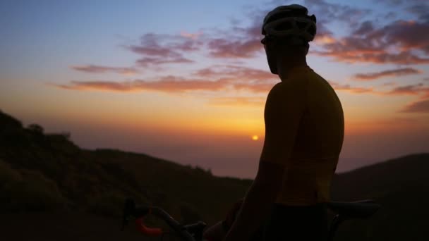 Un hombre en una bicicleta se encuentra en la cima de la montaña y admira la puesta de sol. La cámara se mueve en un Steadicam . — Vídeos de Stock