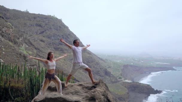 Un homme et une femme debout sur le bord d'une falaise surplombant l'océan lèvent les mains et inhalent l'air marin pendant le yoga — Video