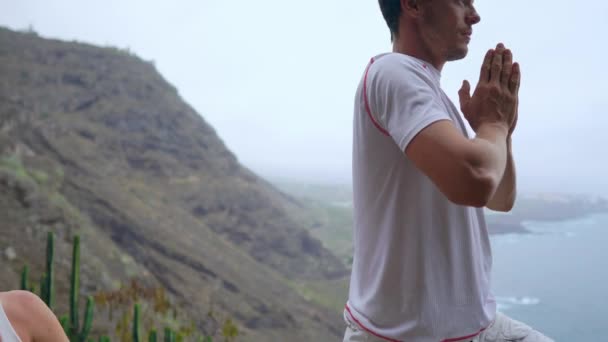 Мужчина и женщина, стоящие на краю скалы с видом на океан, поднимают руки вверх и вдыхают морской воздух во время йоги. — стоковое видео