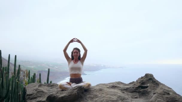 Uma mulher sentada no topo de uma montanha medita e faz um gesto com as mãos Maha Sakal. Contra o oceano e as montanhas verdes — Vídeo de Stock