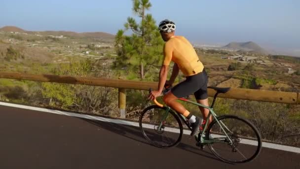 Отслеживание видео, где мужчина-велосипедист поднимается по горной дороге. Человек, занимающийся велоспортом на холмистой дороге в солнечный день . — стоковое видео