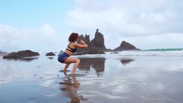 Menina atlética bonita em roupas esportivas na praia do oceano executa agachamentos. Estilo de vida saudável. Aptidão — Vídeo de Stock