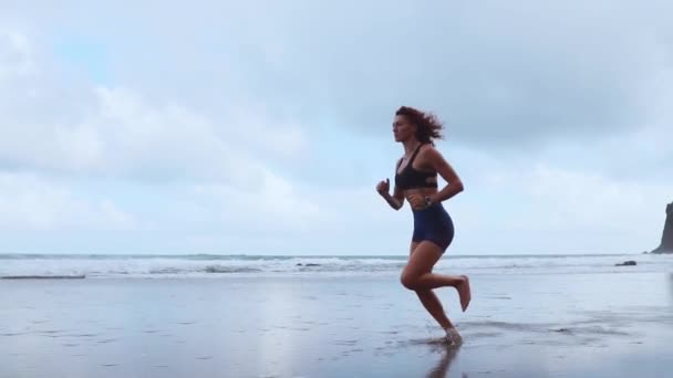 Hermosa chica atleta en ropa deportiva corre a lo largo de la orilla del Océano Atlántico por la mañana. El concepto de un estilo de vida saludable. El físico atlético — Vídeos de Stock