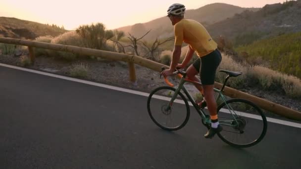 Профессиональный велогонщик в шлеме и спортивном снаряжении едет по горному шоссе на закате в замедленной съемке. Steadicam — стоковое видео