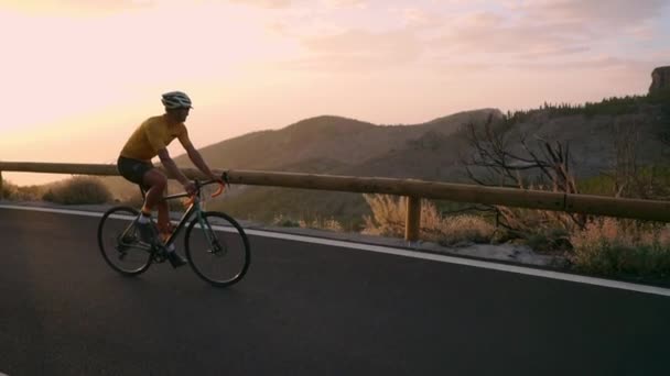 Profesionální cyklista v helmě a sportovní vybavení jezdí na horské silnici při západu slunce ve zpomaleném filmu. Steadicam — Stock video