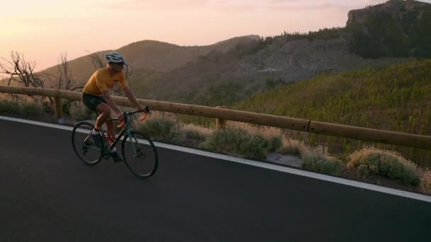 Un ciclista profesional en casco y equipo deportivo monta en una serpentina de montaña al atardecer en dirección al volcán. Steadicam — Vídeos de Stock