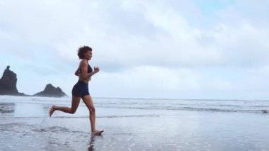 Dağlar ve okyanus ağır çekimde arka plan üzerinde siyah kumlu plajda çalışan spor kadın. Kanarya Adaları
