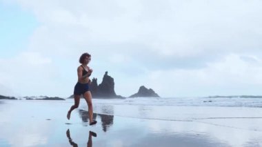 Etkin sportif kadın okyanus sörf zinde ve sağlık tutmak için su havuzu tarafından çalıştırın. Günbatımı siyah kum plaj güneş ile arka plan. Kadın fitness, eğitim ve spor aktivitesi yaz aile tatil koşu.