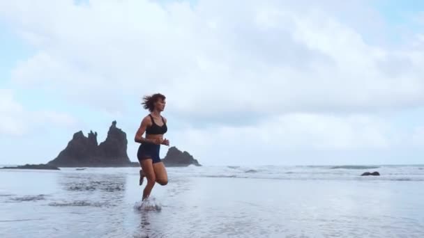Mujer deportiva corriendo en la playa con arena negra en el fondo de las montañas y el océano en cámara lenta . — Vídeo de stock