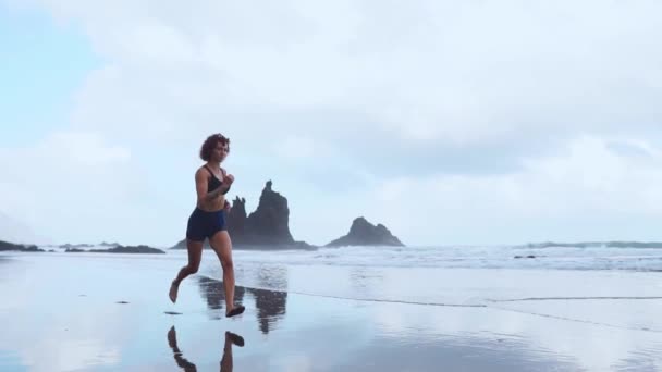 Aktive, sportliche Frau läuft entlang der Meeresbrandung am Wasserpool, um fit und gesund zu bleiben. Sonnenuntergang schwarzer Sandstrand Hintergrund mit Sonne. Frauenfitness, Joggingtraining und sportliche Betätigung im Sommer-Familienurlaub. — Stockvideo