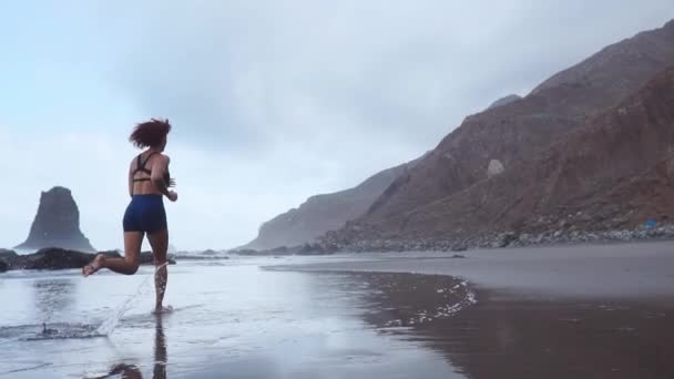 Actieve sportieve vrouw lopen langs de oceaan surf door water zwembad om fit en gezondheid te houden. De achtergrond van de zonsondergang zwarte zand strand met zon. Vrouw fitness, joggen opleiding en sport activiteit op zomervakantie met het gezin. — Stockvideo