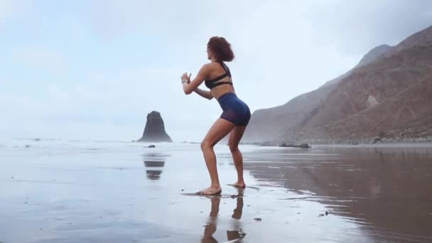 Una mujer hace sentadillas en las orillas del océano Atlántico. El concepto de un estilo de vida saludable. Practicar deportes en Canarias — Vídeo de stock